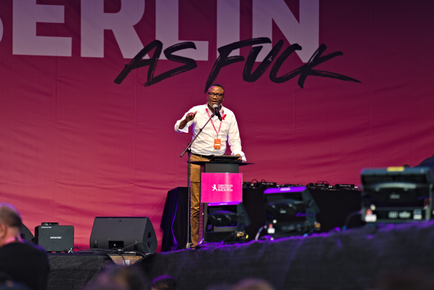 Ein Mann in einem weißen Hemd und braunen Hosen steht auf einer Bühne und hält ein Mikrofon in der Hand, hinter ihm eine pinkfarbene Fahne mit der Aufschrift "Berlin as Fuck" und Lautsprecher und andere Gegenstände auf der Bühne.