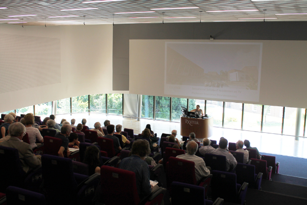 Ein Seminarraum in einer Universität mit vielen Menschen, die aufmerksam einer Präsentation an der Front zugewandt sind, wo eine Person an einem Tisch steht und den Inhalt erklärt, mit Glasfenstern und Blicken auf Bäume und Gebäude im Hintergrund.