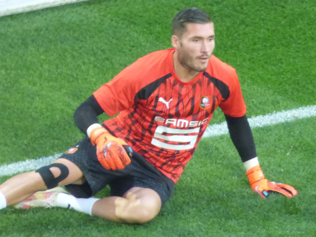 Ein Fußballspieler sitzt auf dem Boden mit Händen auf den Knien, trägt ein rotes T-Shirt, schwarze Shorts, weiße Socken und orangefarbene Baseball-Handschuhe.