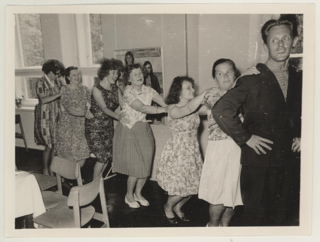 Eine Gruppe von Menschen tanzt in einem Raum im Stil der 1950er Jahre mit Stühlen, einem Tisch mit einer weißen Decke, einem Foto auf der Wand und Fenstern im Hintergrund.