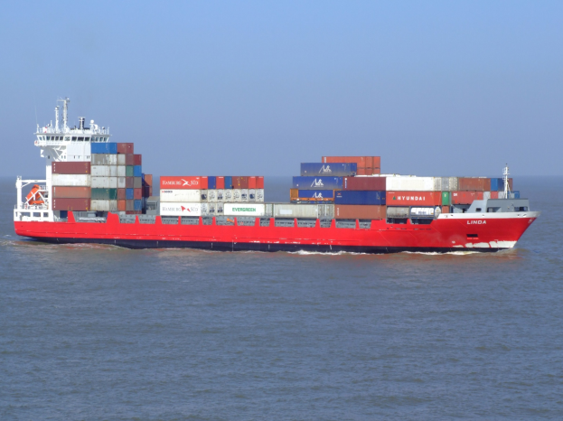 Ein großes rotes und weißes Containerschiff mit vielen Containern auf seinem Deck segelt auf dem Meer unter einem klaren blauen Himmel.