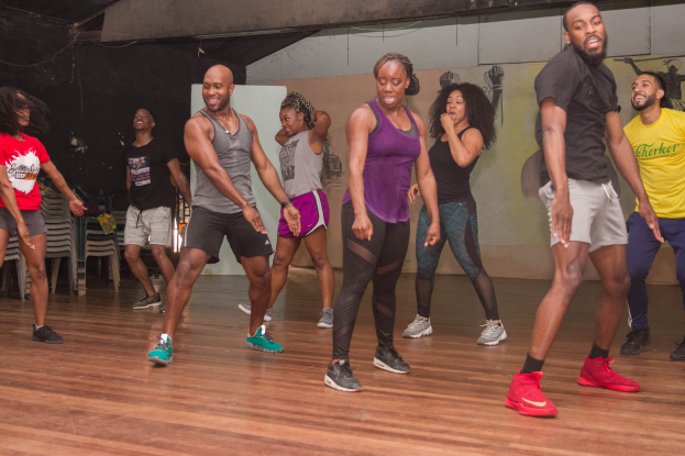 Eine Gruppe von Menschen, die in einem Tanzstudio tanzen, tragen Turnschuhe, mit einer bemalten Wand, Stühlen und anderen Gegenständen im Hintergrund, Deckenleuchten darüber und der Boden sichtbar darunter.