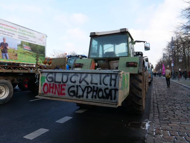 Ein Traktor mit Graffiti auf der Rückseite fährt eine Straße entlang, umgeben von Menschen in verschiedenenfarbigen Kleidern, mit Bäumen und Polen am Straßenrand und einem Himmel im Hintergrund.