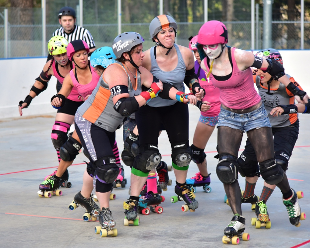 Mehrere Frauen mit Helmen und Rollschuhen sind Rollschuhlaufen, scheinbar gegenseitig schiebend in der Nähe eines Metallzauns, mit einem Mann und Bäumen im Hintergrund.