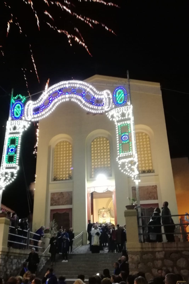 Eine Gruppe von Menschen steht vor einem Gebäude mit einem beleuchteten Torbogen, Treppen mit Geländern und Topfpflanzen, während Feuerwerk den Nachthimmel erhellt.