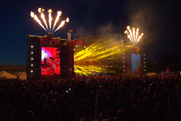Eine große Menschenmenge auf einem Musikfestival mit einer beleuchteten Bühne, die Musiker, Banner, Zelte links und Feuerwerke am dunklen Himmel zeigt.