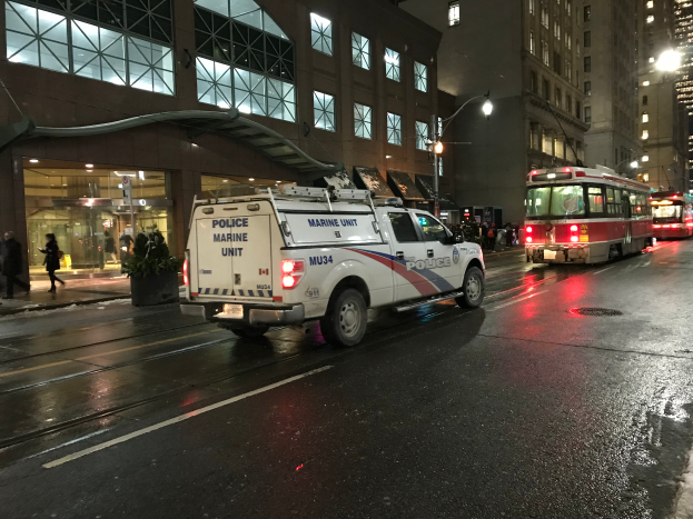 Zwei Männer kehren eine Straße mit Besen, umgeben von Gebäuden, Menschen, Pfählen, Brettern und anderen Gegenständen, mit Text am unteren Bildrand.