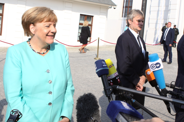 Bundeskanzlerin Angela Merkel beim Sprechen mit Reportern vor einem Gebäude mit einer Gruppe von Menschen und Medienausrüstung, unter einem bewölkten Himmel.