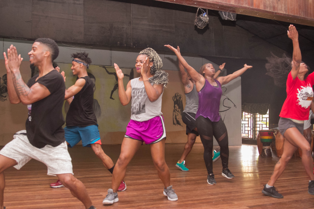 Eine Gruppe von Menschen tanzt in einem Tanzstudio mit einem Gemälde an der Wand und einem Glasfenster auf der rechten Seite, unter Deckenlampen, während einer Zumba-Stunde.
