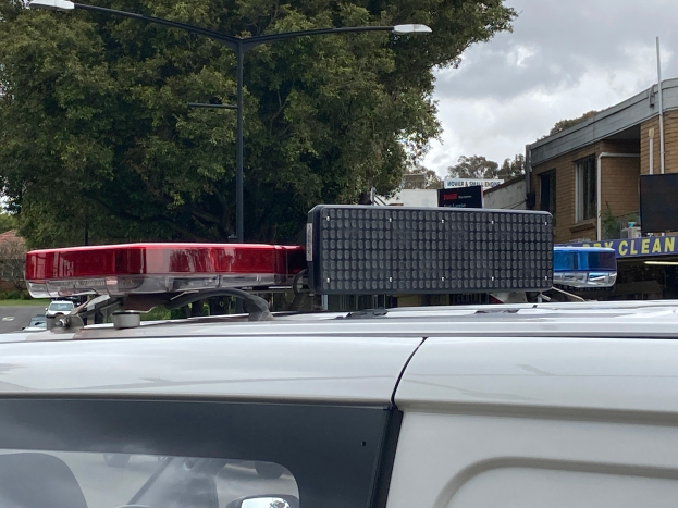 Ein Polizeiwagen mit eingeschaltetem Blaulicht steht am Straßenrand umgeben von Gebäuden, Bäumen, Pfählen und Texttafeln, mit einem bewölkten Himmel im Hintergrund, wahrscheinlich nach einem tödlichen Schulschießen in Brisbane.
