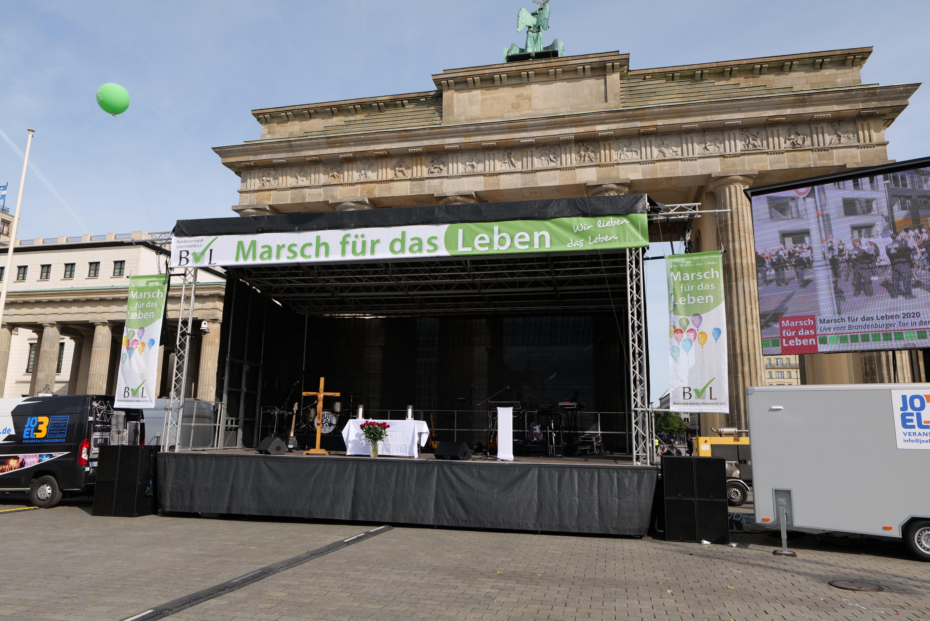 Bühne vor dem Brandenburger Tor mit einem Tisch, Fahnen, Lautsprechern, Fahrzeugen, Gebäuden, einer Statue, einer Flagge und Wolken am Himmel.