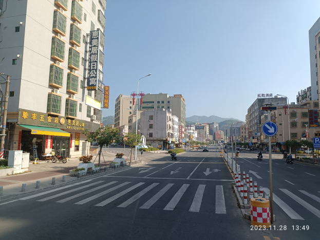 Stadtstraße mit Gebäuden, Straßeninfrastruktur, Fahrzeugen, Bäumen, Hügeln und Himmel im Hintergrund.