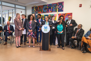 Eine Gruppe von Menschen, die vor einem Podium mit einem Mikrofon und einer Tafel mit der Aufschrift "Sancho City Councilman Joins City Council" stehen und sitzen, mit Flaggen, einer Tafel, einem Feuerlöscher und Deckenleuchten an der Rückwand.