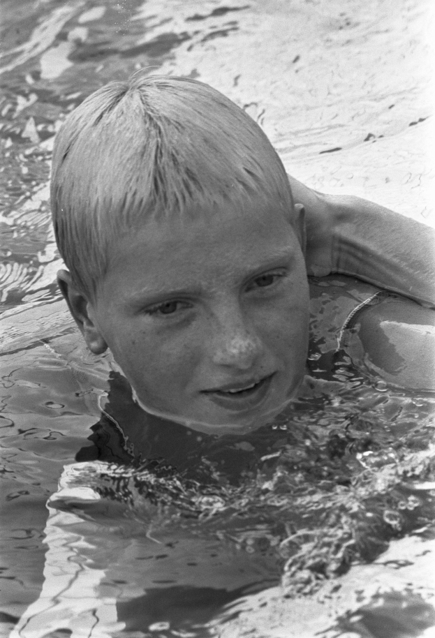 Ein junger Junge schwimmt in einem Pool mit ausgestreckten Armen, das Gesicht von der Sonne beleuchtet, während er durch das Wasser gleitet, die Augen geschlossen und das Haar nass.
