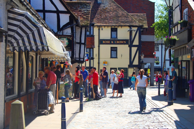 Eine Straße mit Menschen auf der linken Seite neben einem Handwagen und einem Mann und einer Frau auf der rechten Seite neben einem lilafarbenen Müllcontainer, mit Gebäuden, Bäumen und Blumentöpfen im Hintergrund.