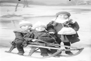 Drei Kinder in Mänteln und Mützen sitzen auf einem hölzernen Schlitten im Schnee, mit Bäumen im Hintergrund.