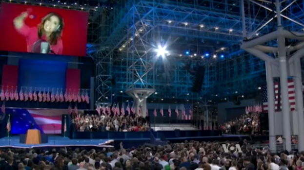 Eine große Menschenmenge sitzt vor einer Bühne mit einem Rednerpult, Mikrofon, Flaggen, einem Bildschirm mit einer Frau und Beleuchtungsausrüstung, wahrscheinlich bei einer Präsidentschaftsdebatte.