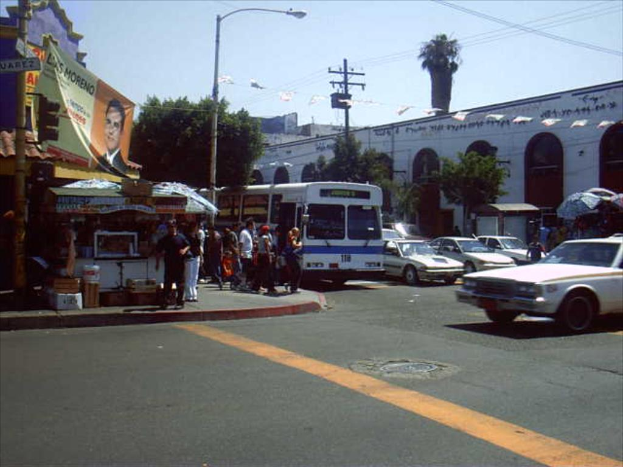 Eine belebte Straßenszene mit zahlreichen Fahrzeugen, Gebäuden, einem Stand, Bäumen und einem Himmel darüber.