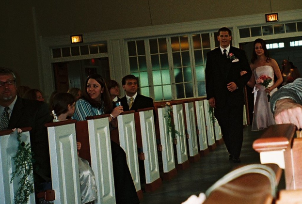 Eine Gruppe von Menschen sitzt auf einer Bank, während eine Braut und ein Bräutigam den Weg entlang auf sie zukommen, die Braut hält einen Blumenstrauß, mit Laternen und einem Fenster im Hintergrund, was auf eine Kirchenatmosphäre hindeutet.