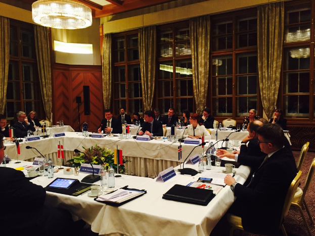 Menschen an einem Tisch mit verschiedenen Büromaterialien, darunter Ordner, Papiere, Getränke und elektronische Geräte, in einem Konferenzraum mit roter Wand, grünen Vorhängen und sichtbarem Dach.