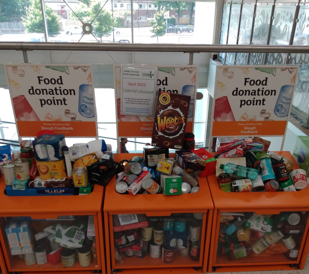 Orange Schübe mit Lebensmittelspenden wie Flaschen, Dosen und Kartons, mit Informationsschildern und einem Blick durch Glasfenster auf Fahrzeuge, Bäume, Gebäude und Himmel.