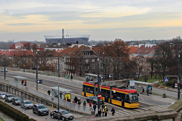 Eine gelbe und schwarze Straßenbahn fährt auf einer von hohen Gebäuden gesäumten Stadtstraße, umgeben von Autos, Fußgängern, Laternen, Schildern, Bäumen und Gras, mit bewölktem Himmel im Hintergrund.