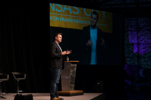 Eine Person in einem schwarzen Hemd hält einen Vortrag, steht neben einem Tisch mit einer Flasche, einem Stuhl dahinter und einem Projektor, der links von ihr etwas anzeigt, mit einem schwarzen Vorhang im Hintergrund.