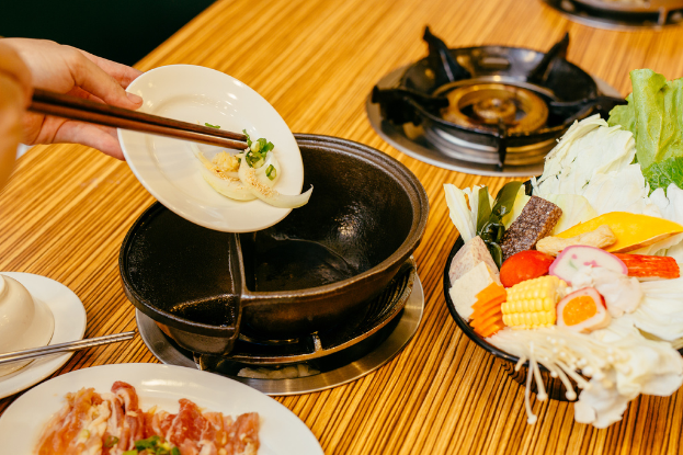 Eine Person hält einen Teller in den Händen in der Nähe eines Tisches mit einem Gasherd, einer Pfanne, einer Schüssel mit Obst und einem Teller mit Essen.