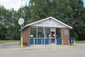 Ein kleines United States Post Office Gebäude in Lake George, Michigan, mit Glasfenstern und -türen, einer Namensplakette an der Wand, einem Pfahl mit einer angebrachten Plakette, gelben Pfählen, einem blauen Müllcontainer, Gras, Bäumen und einem bewölkten Himmel im Hintergrund.