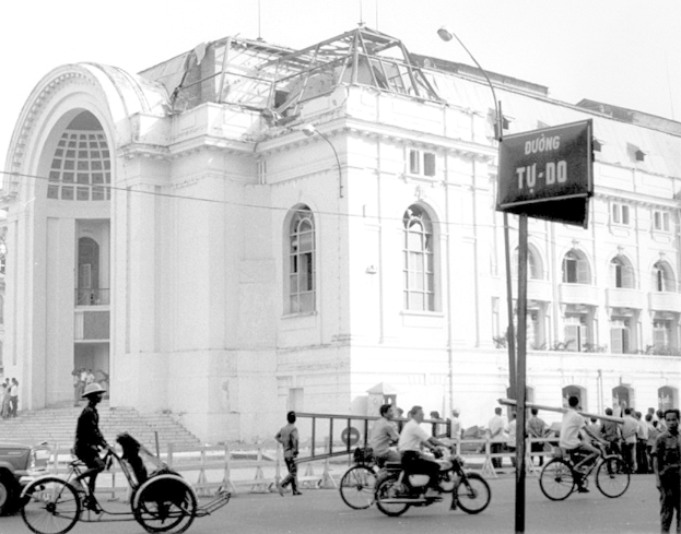 Ein Gebäude mit Menschen, die Fahrzeuge auf der Straße fahren, Straßenlaternen und einige Personen, die auf der Straße stehen.
