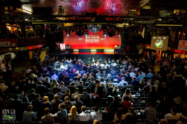 Eine große Menschenmenge sitzt auf Stühlen vor einer Bühne mit Bildschirmen und Bannern, die von Scheinwerfern beleuchtet wird und von Geländern umgeben ist, und blickt erwartungsvoll auf den Beginn eines Online-Marketing-Rockstars-Events, mit einem Wasserzeichen in der unteren linken Ecke.