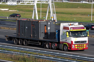 Ein Lastwagen mit einem großen Container fährt auf einer Autobahn, mit anderen Fahrzeugen, Pfählen, Bäumen, Gebäuden und einem klaren blauen Himmel im Hintergrund.