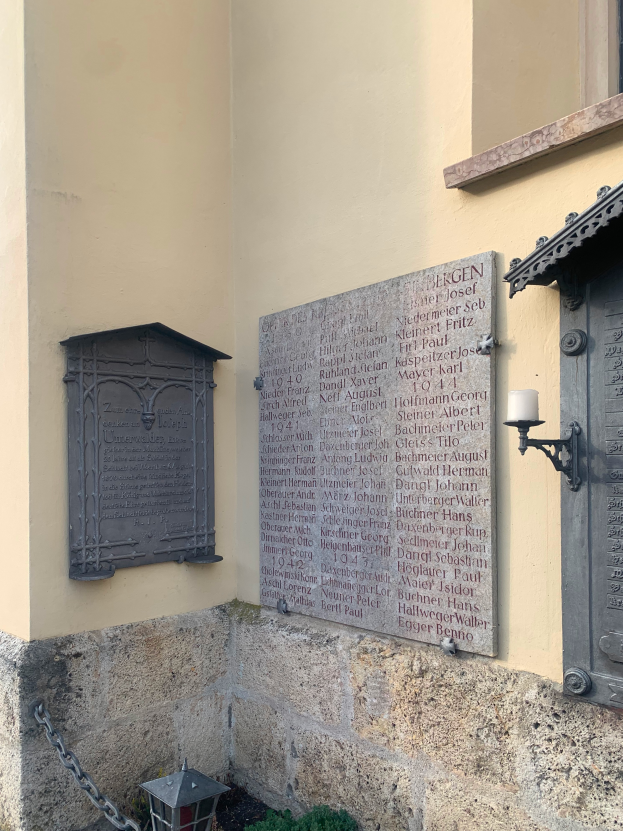 Ein Gebäude mit einer Gedenktafel, einem Fenster, einer Tür, einem Licht, einer Kette und einigen Pflanzen, die Holocaust-Opfer und Kriegsopfer gedenken.
