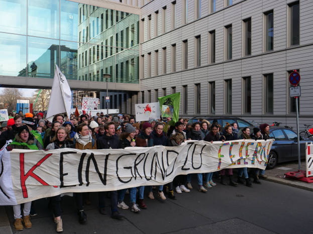 Eine Gruppe von Menschen, die auf der Straße stehen und ein Transparent mit der Aufschrift "Kein Grind Lo Feuer" tragen, mit Gebäuden, Bäumen, Masten, Schildern, Fahrzeugen und einem klaren blauen Himmel im Hintergrund.