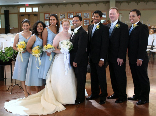 Eine Frau in einem weißen Kleid mit einem Blumenstrauß steht in der Mitte, umgeben von Frauen in blauen Kleidern mit Blumenstrauß und Männern in schwarzen Anzügen, die alle lächeln und drinnen mit verschiedenen Gegenständen und Dekorationen im Hintergrund stehen.