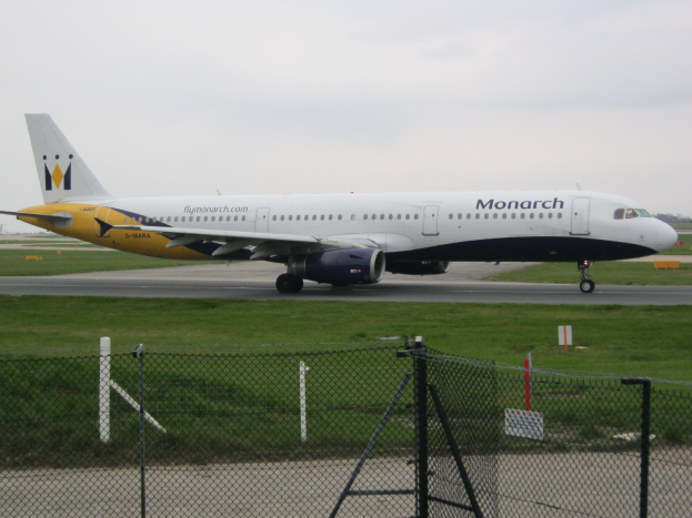 Eine Monarch Airlines Airbus A320-200 auf einer Rollbahn, umgeben von Gras und einem Zaun mit Pfählen und Brettern, mit Bäumen und einem klaren blauen Himmel im Hintergrund.