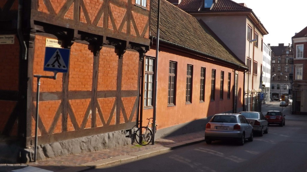 Fahrzeuge auf der Stra├če mit Geb├Ąuden, Laternen, Schildermasten, Fenstern und D├Ąchern im Hintergrund; ein Fahrrad ist auf dem Gehweg in der N├Ąhe einer Stange abgestellt.