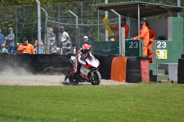 Eine Person auf einem Motorrad fährt über ein grasbewachsenes Feld, mit Zuschauern in Mützen und Schutzbrillen, einem Maschendrahtzaun, Pfosten, Bäumen, Treppen mit Geländern und einem Schuppen mit Nummern und Texttafeln im Hintergrund.