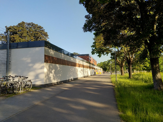 Ein Fahrradweg mit mehreren parkenden Fahrrädern am Rand, umgeben von Gras und Bäumen, mit Gebäuden und einem klaren blauen Himmel im Hintergrund.