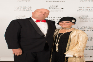 Ein Mann in einem schwarzen Anzug mit weißem Hemd und schwarzer Fliege steht neben einer Frau in einem eleganten Kleid mit einem gemusterten Schultertuch auf einem roten Teppich, mit einem weißen Banner mit Text im Hintergrund.