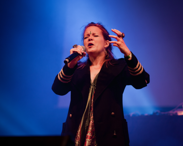 Eine Frau in einem schwarzen Kleid singt auf der Bühne in ein Mikrofon, ihre Augen geschlossen und von einem blauen Licht erhellt.