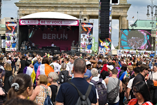 Eine große Menschenmenge steht vor dem Brandenburger Tor, in der Mitte eine Bühne mit Musikern und Bannern, ein großer Bildschirm rechts, Leuchtpfähle links und ein bewölkter Himmel im Hintergrund.