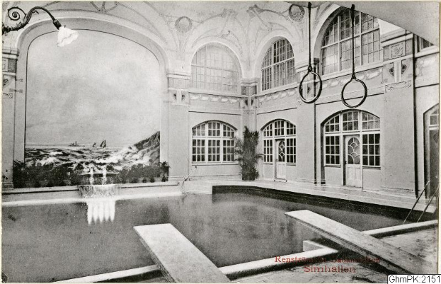 Ein altes Schwarz-Weiß-Foto eines großen Hallenbades in einem Gebäude, umgeben von einer Wand mit Glasfenstern und -türen, mit Pflanzen und einer Deckenleuchte und Text unten, der 'Rensburg, Deutschland - Schwimmbad' lautet.