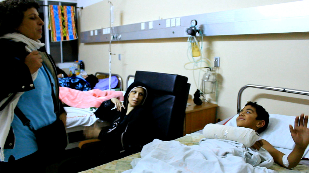 Ein Krankenhauszimmer mit mehreren Betten, auf denen Patienten liegen, Infusionsflaschen auf Ständern zwischen den Betten und eine Frau mit einer Tasche in der Nähe eines Patienten.