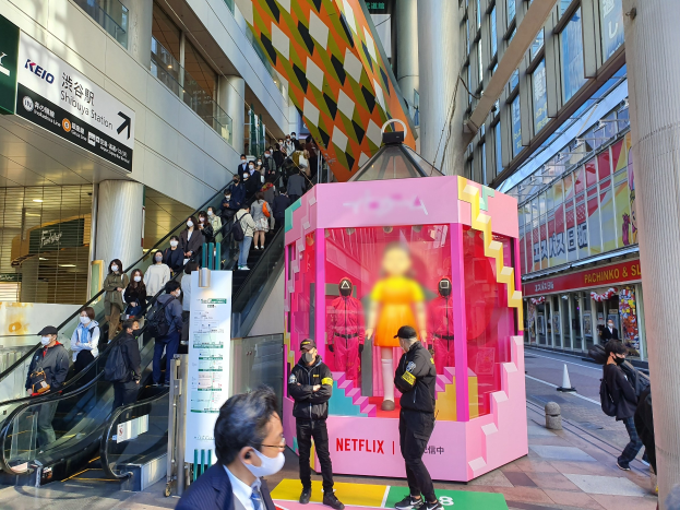 Eine Gruppe von Menschen steht vor einem Gebäude mit Glaswänden, Säulen und Texttafeln, einige tragen Masken, während hinter ihnen eine Rolltreppe mit Menschen nach oben führt; eine pinkfarbene Box in der Mitte zeigt Schaufensterpuppen und trägt den Text "Netflix's neuer Netflix-Stand in Tokyo."
