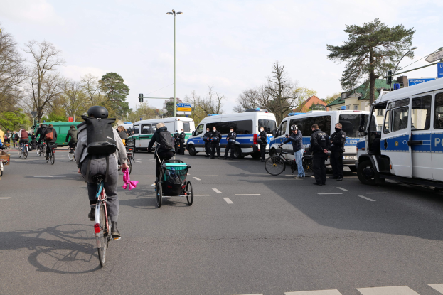 Eine Gruppe von Menschen auf Fahrrädern auf einer Straße mit Polizeiwagen und anderen Fahrzeugen, einige tragen Helme und haben Taschen oder schieben Kinderwagen, vor dem Hintergrund von Bäumen, Gebäuden und Verkehrsinfrastruktur.