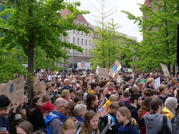 Eine große Menge von Menschen protestiert vor einem Gebäude in Berlin, mit Schildern, Bäumen, Fahrzeugen und einem Lautsprecher.