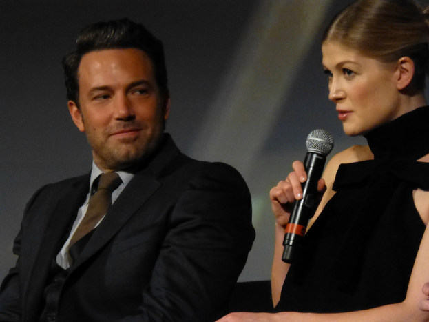 Eine Frau hält ein Mikrofon und spricht, während ein Mann in einem Blazer und Krawatte sie ansieht und lächelt.