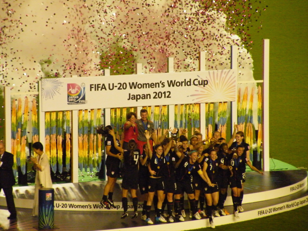 Gruppe von Frauenfußballspielerinnen in schwarzen Uniformen, die auf einer Bühne umgeben von einer Menge feiern, mit einem Hintergrund aus Text, Bildern und Dekorationen auf Gras.