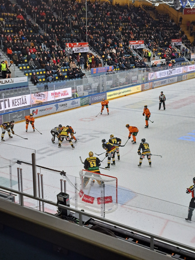 Eine Gruppe von Menschen, die Hockey auf einem Eisstadion spielt, mit einem Netz, einer Umzäunung, sitzenden Zuschauern und beleuchteten Lichtern.